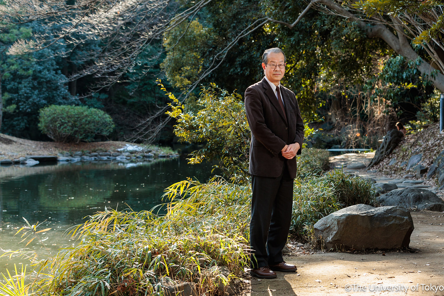 Photograph of Professor Yutaka Michida, wearing a dark suit, standing next to Sanshiro pond at the University of Tokyo on a sunny day.