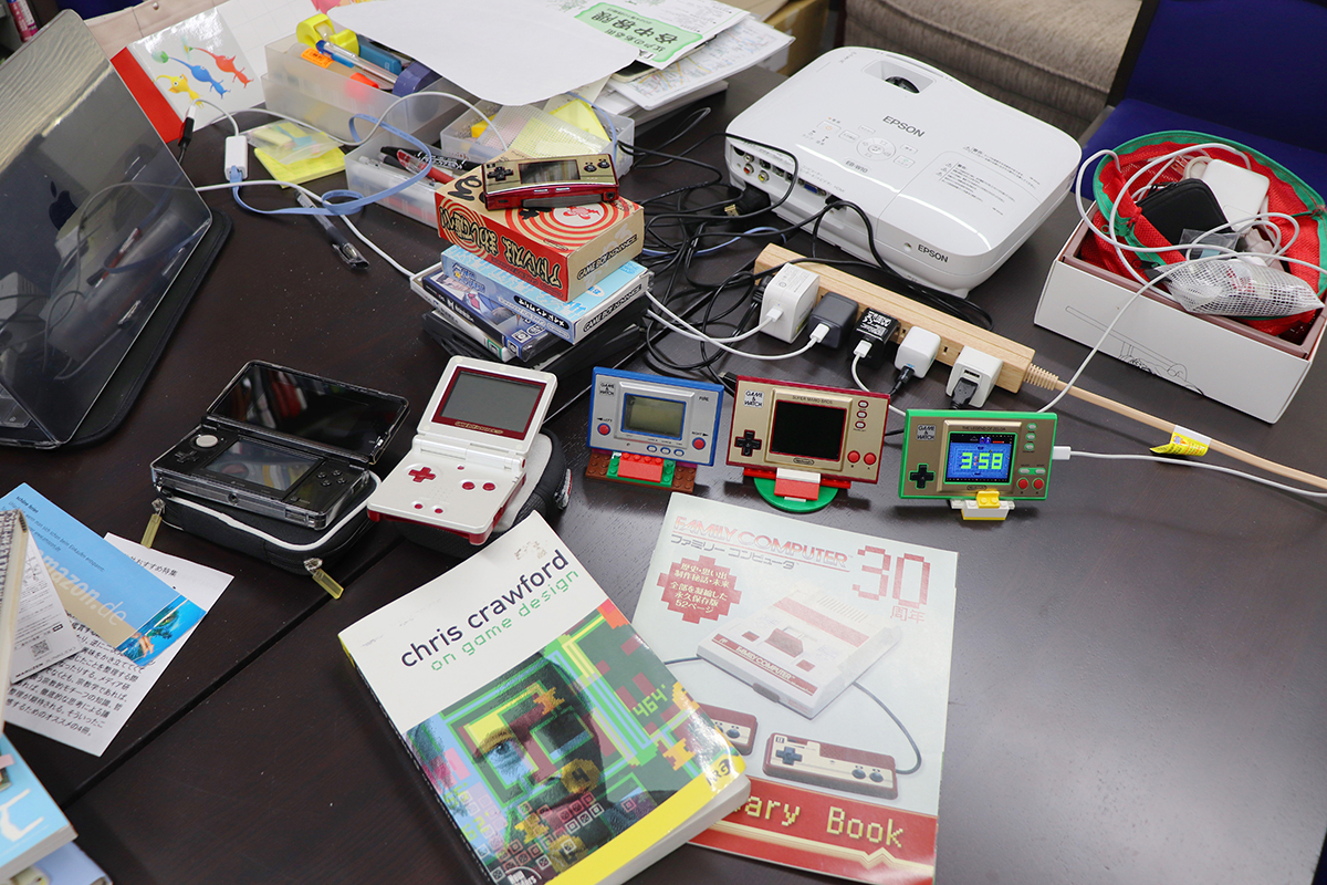 研究室の机の上に「ゲーム＆ウオッチ」「ゲームボーイ」などのデバイスや関連図書が置かれている